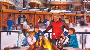 A group of people around a campfire in a stone fire pit with snow on the ground, surrounded by buildings with wooden railings.
