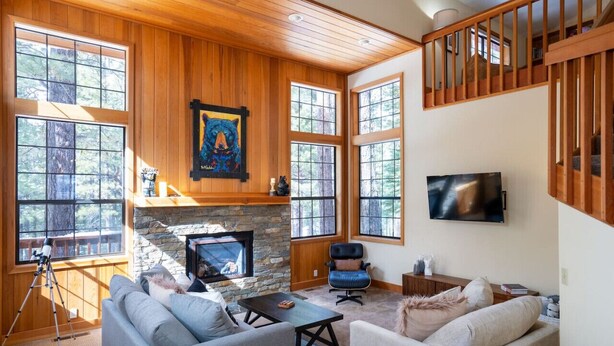 A cozy living room with a stone fireplace, large windows, a mounted TV, and a wooden staircase.