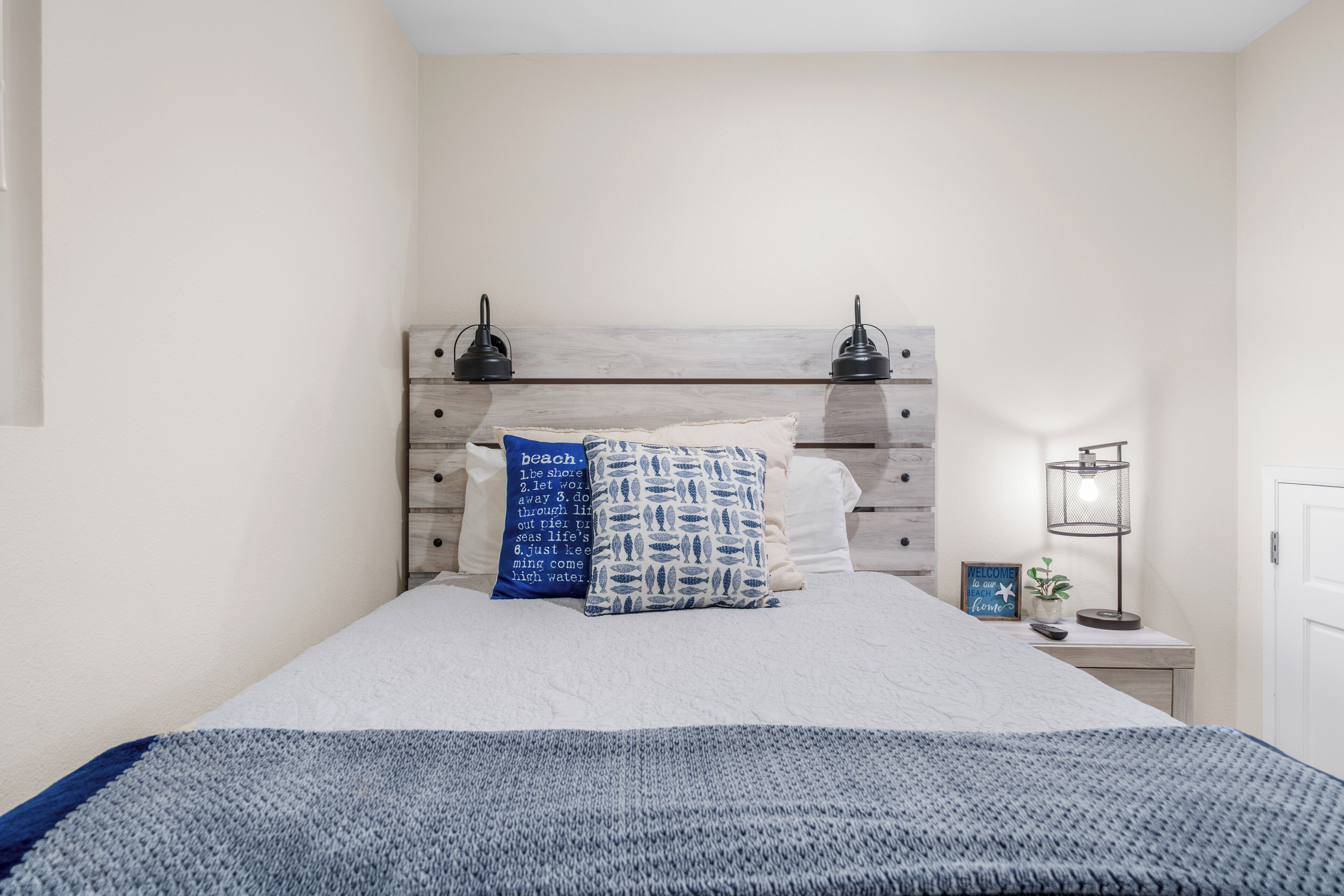 A neatly made bed with a blue and white patterned pillow, a wooden headboard, and a bedside table with a lamp and a small plant.