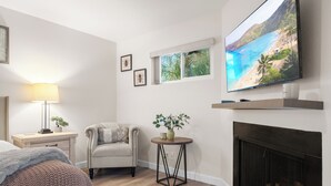 Een slaapkamer met een bed, een nachtkastje met lamp, een stoel, een kleine tafel met plant en een open haard, en een televisie aan de muur bevestigd.