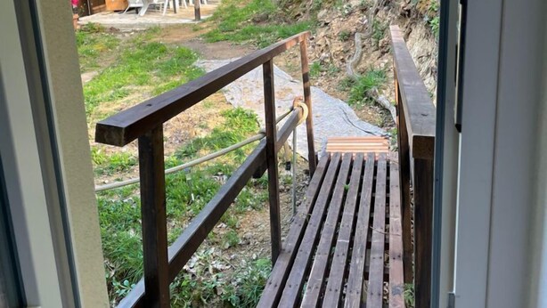 Eine Holzterrasse mit Metallgeländer, die zu einem überdachten Außenbereich mit roten Vorhängen und einem Tisch führt.