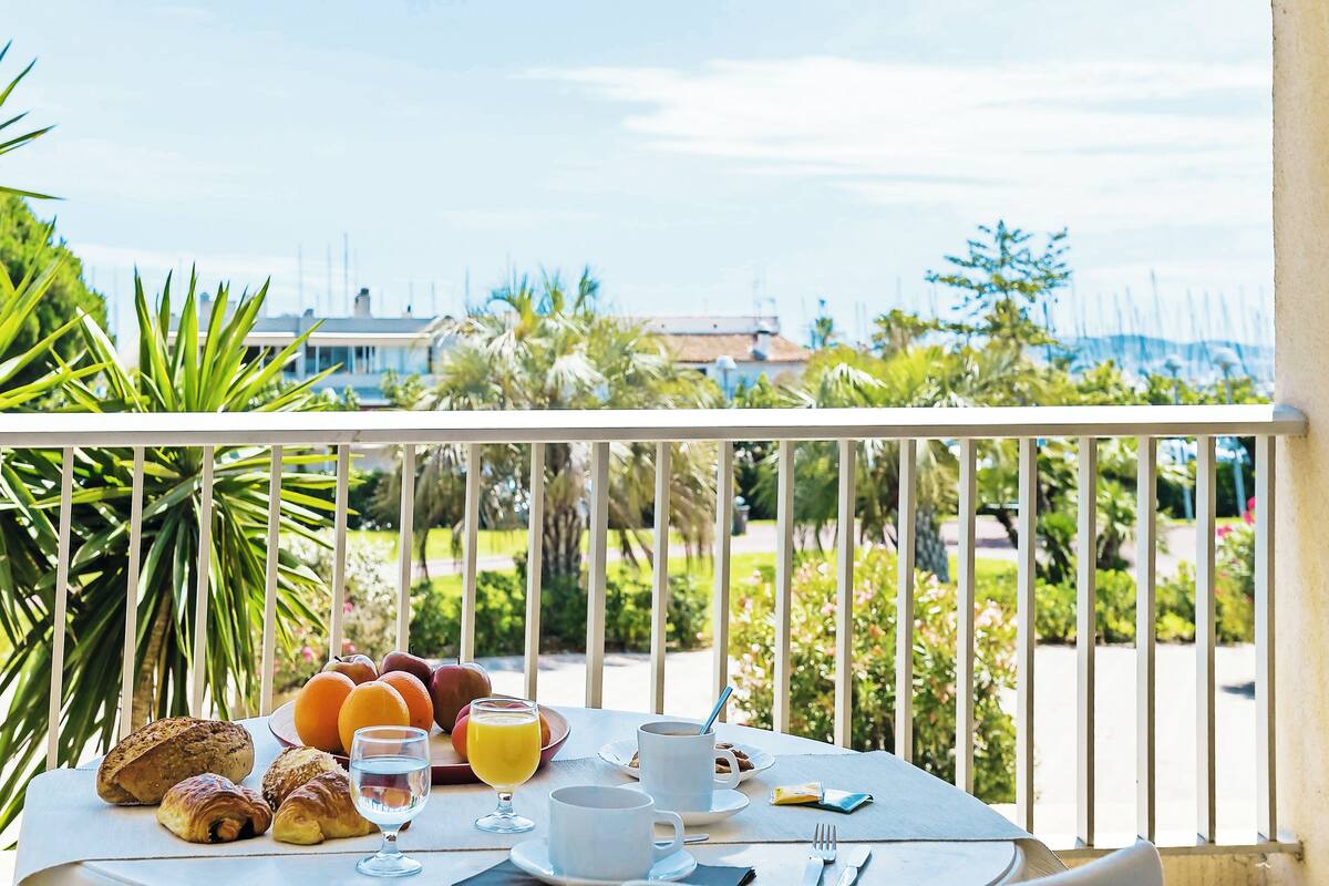 Ein Balkon mit Frühstückstisch, auf dem Croissants, Orangensaft und Kaffee bereitstehen, mit Blick auf einen Garten und Gebäude.