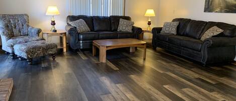 Ein Wohnzimmer mit dunklem Holzboden, zwei schwarzen Ledersofas mit gemusterten Kissen, einem Holztisch und zwei Lampen auf Beistelltischen.