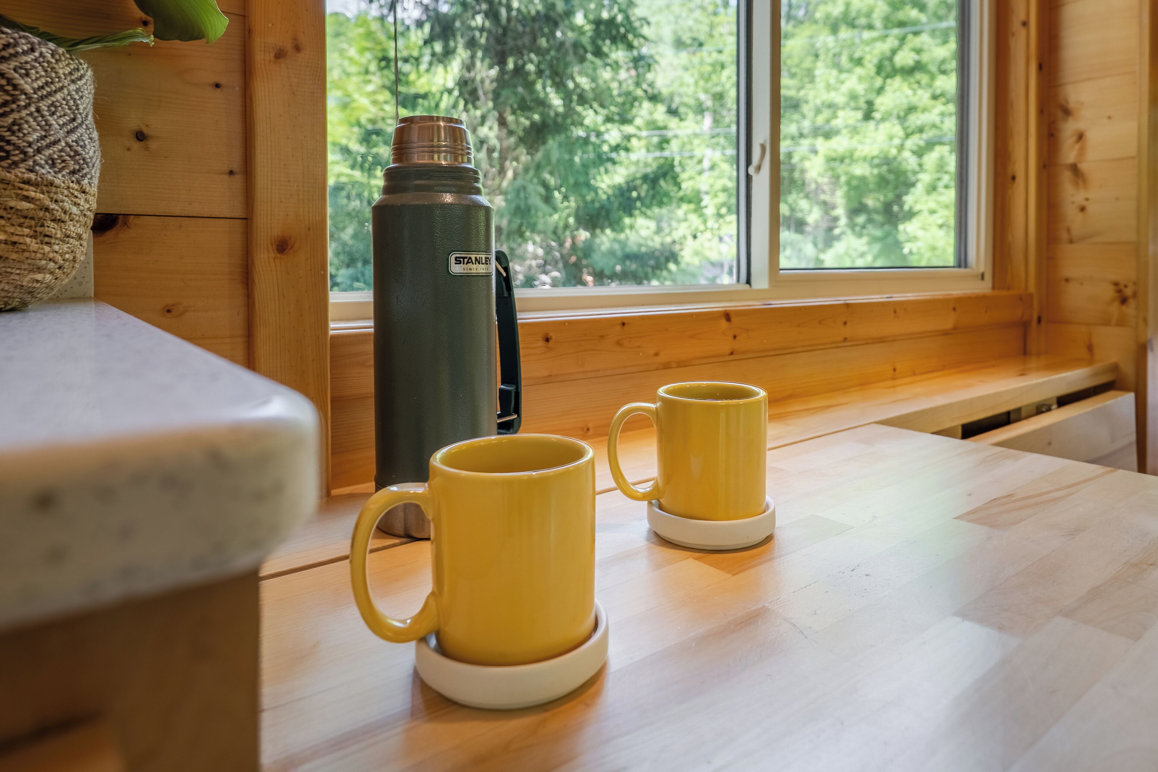 Un intérieur en bois avec une fenêtre, une gourde noire de la marque Stanley et deux tasses jaunes posées sur une table en bois.