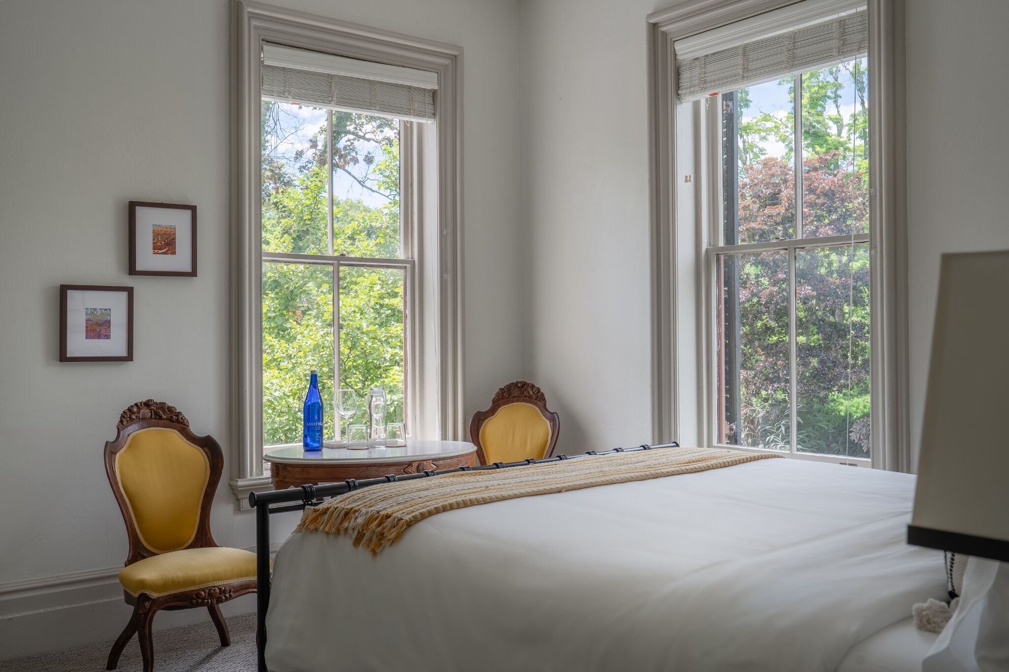Habitación de hotel con una cama grande, dos sillas, una mesita con una botella y cuadros enmarcados en la pared.