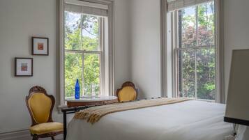 Habitación de hotel con una cama grande, dos sillas, una mesita con una botella y cuadros enmarcados en la pared.