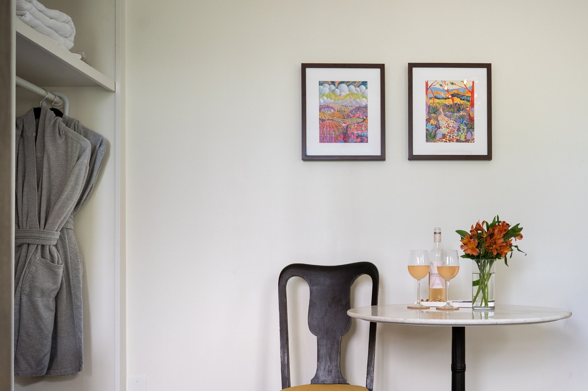Une pièce comprenant un lit, une robe suspendue, deux œuvres d’art encadrées, une chaise, une table avec des verres et des fleurs, et une bouteille.