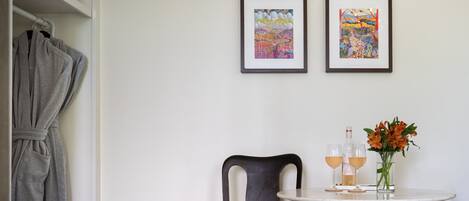 Une pièce comprenant un lit, une robe suspendue, deux œuvres d’art encadrées, une chaise, une table avec des verres et des fleurs, et une bouteille.