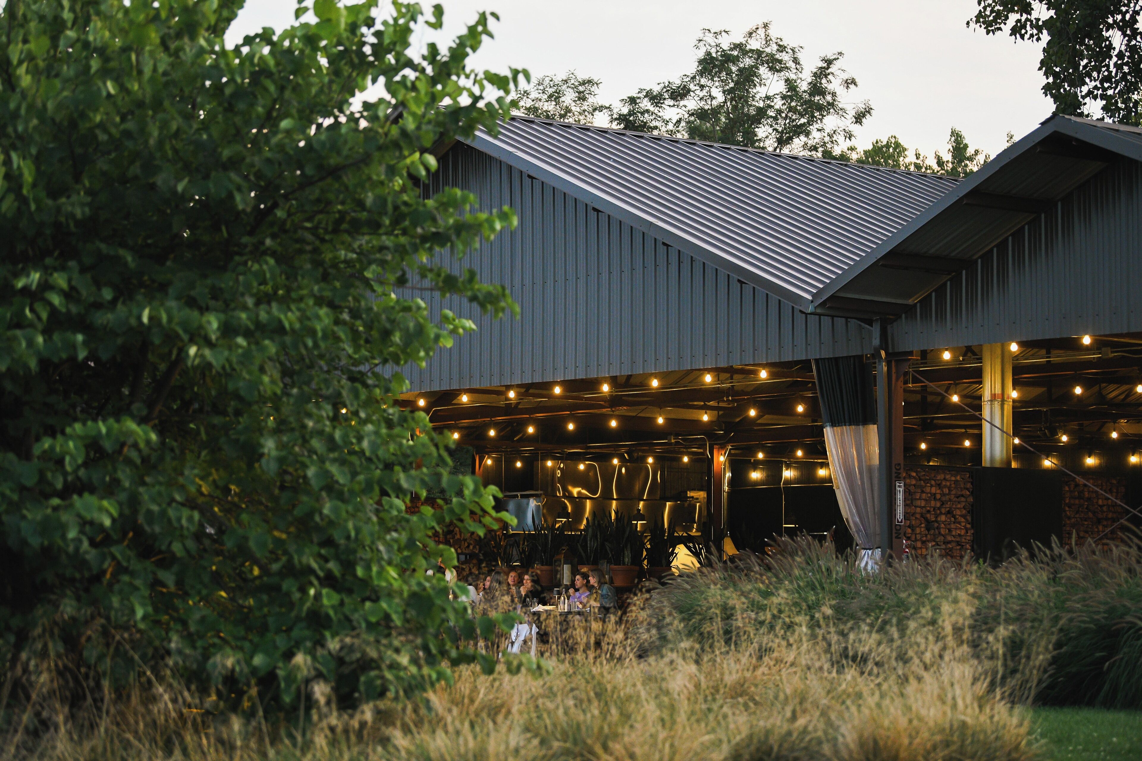An outdoor dining area with tables and chairs, surrounded by greenery and a building with a metal roof.