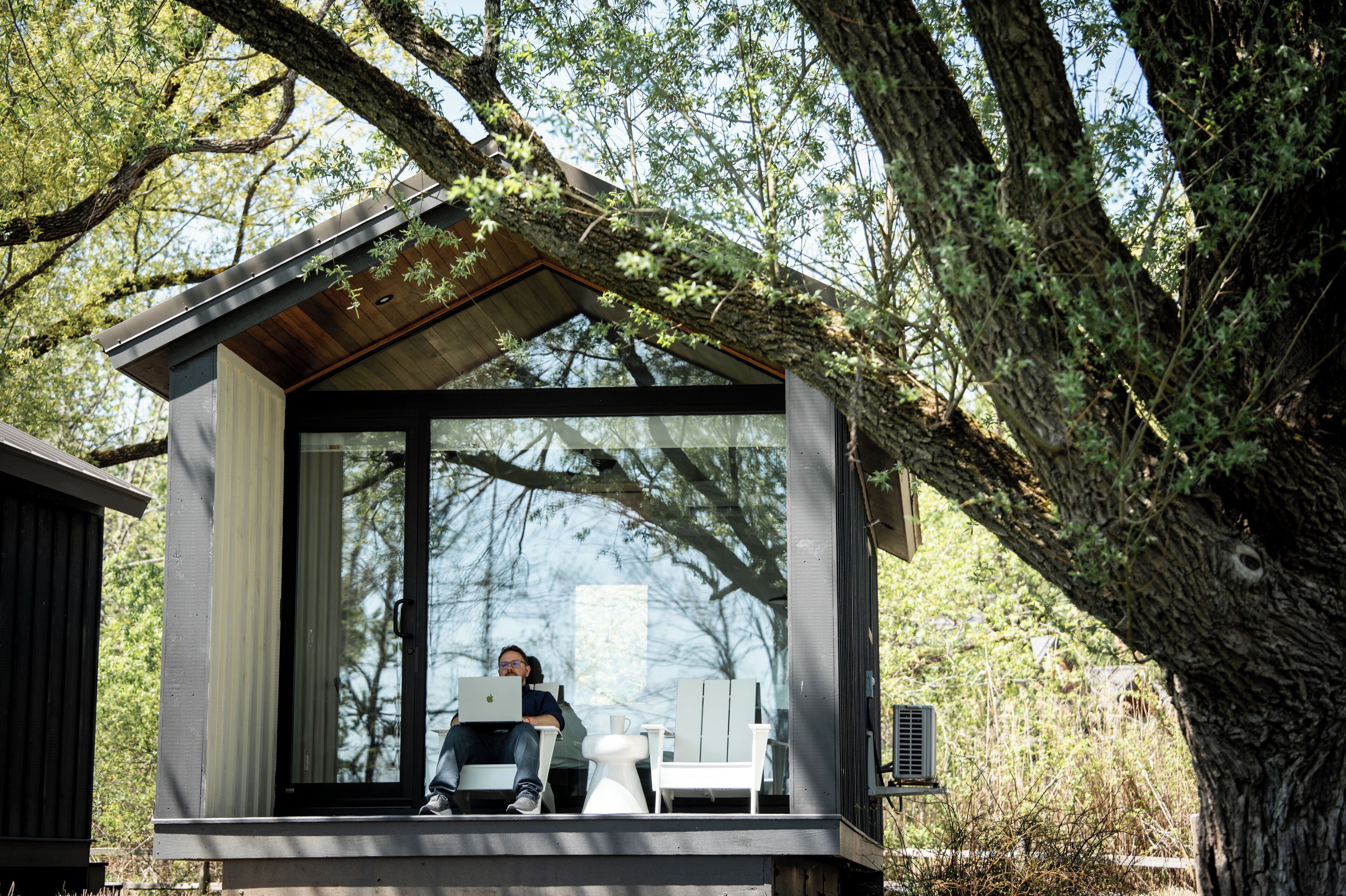 A modern cabin with a glass door, white furniture, and a large tree in the background.