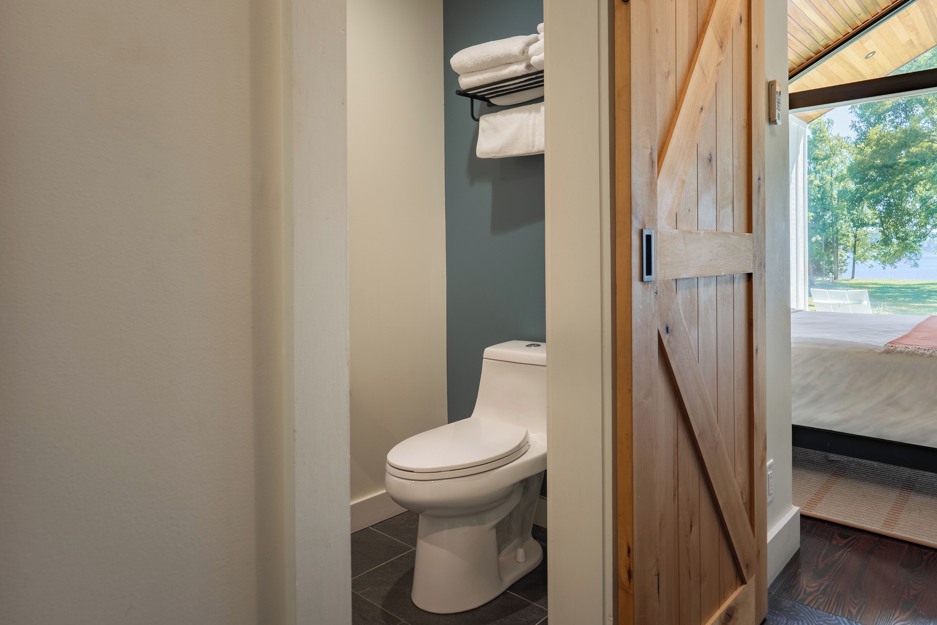 A bathroom with a toilet, a towel rack, and a view of a bed and a window.
