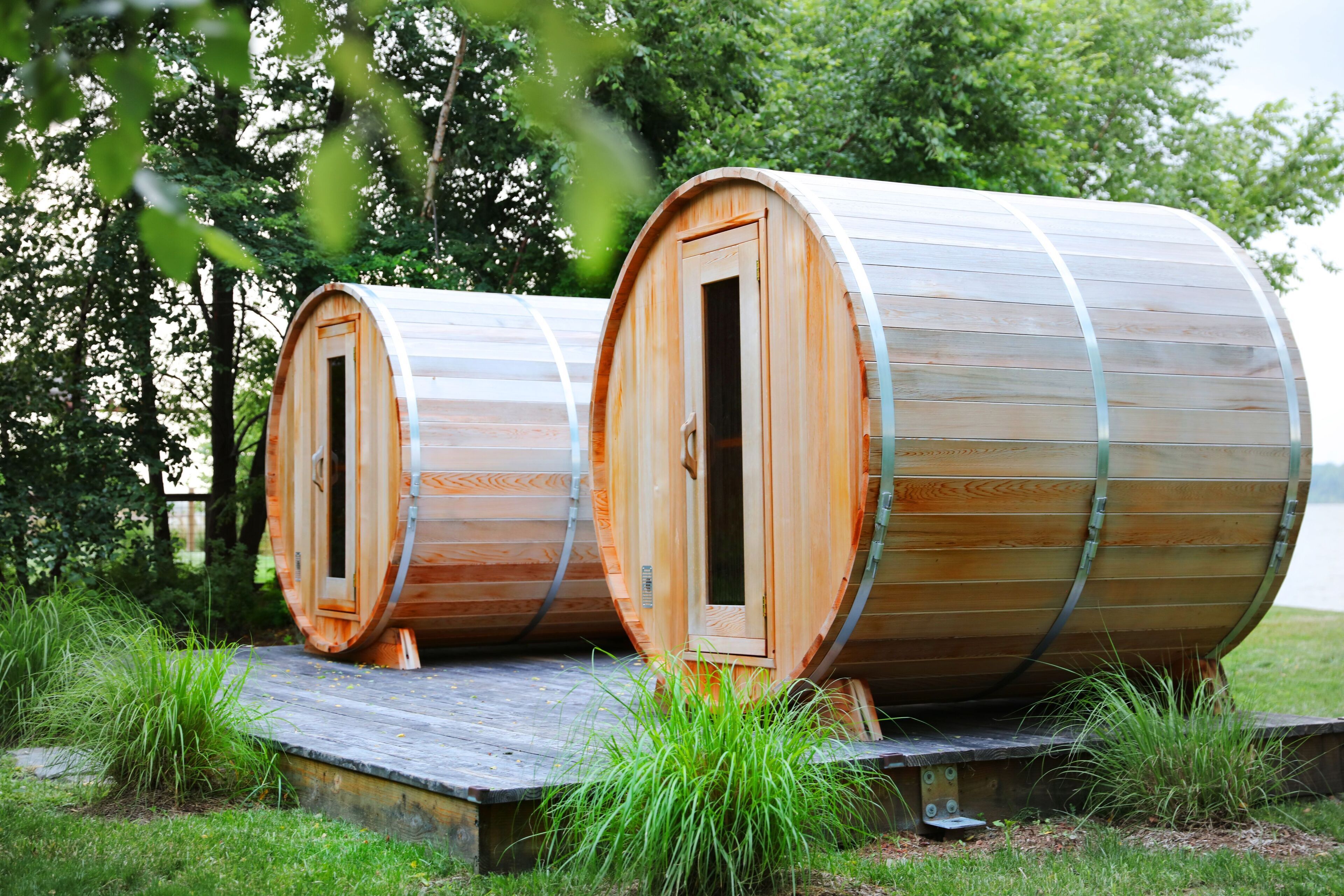 Two wooden barrel-shaped cabins with doors, set on a wooden platform in a grassy area with trees in the background.