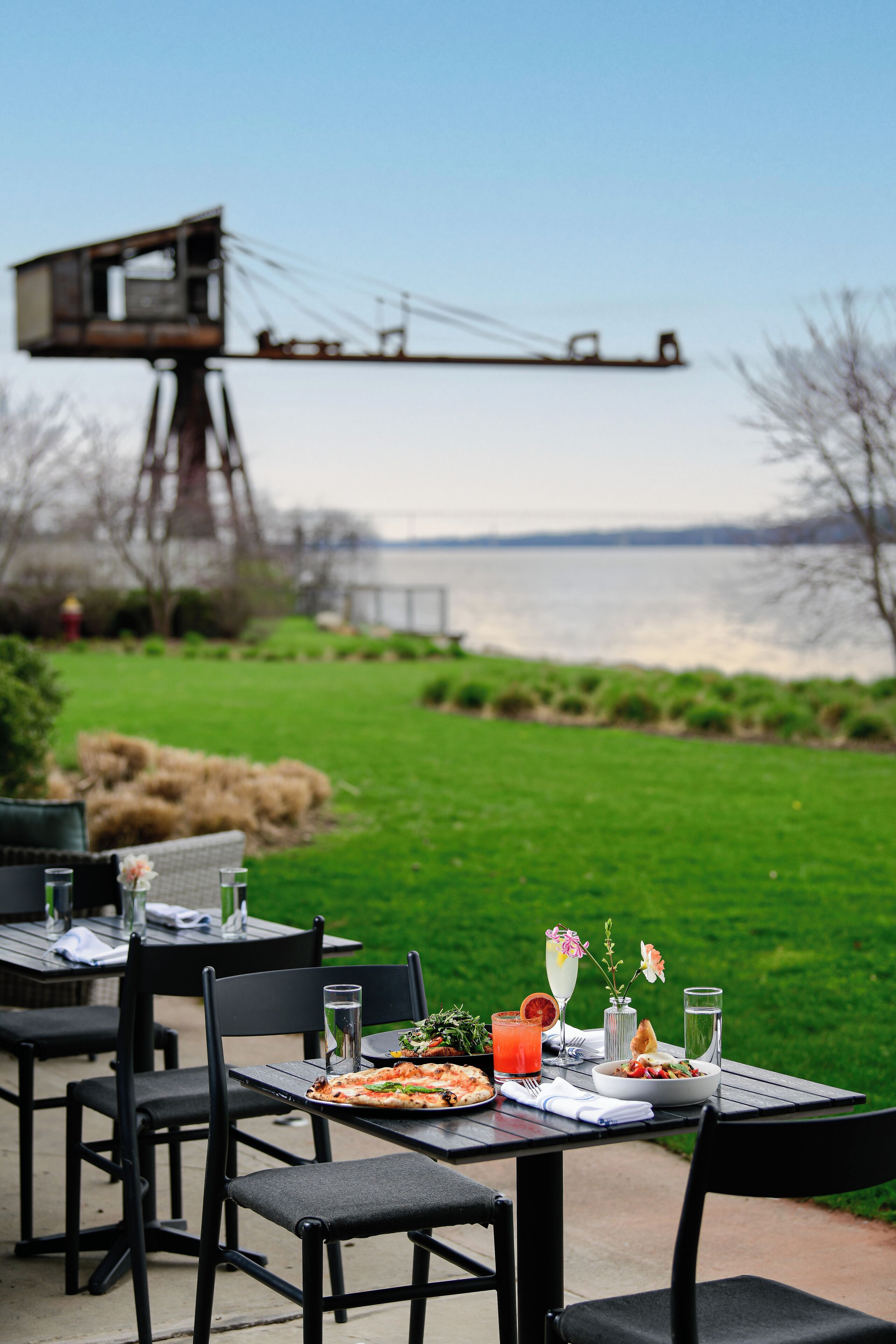 Un espacio al aire libre con mesas y sillas para comer, una pizza, una ensalada y una bebida, con vistas a un cuerpo de agua y una estructura distante.