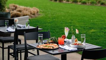 Un espacio al aire libre con mesas y sillas para comer, una pizza, una ensalada y una bebida, con vistas a un cuerpo de agua y una estructura distante.
