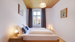 Un dormitorio con techo de madera, un ventanal con cortinas, una cama con ropa blanca y dos lámparas de noche.