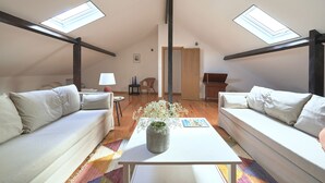 A cozy loft bedroom with a white sofa, a small table with a vase, and a colorful rug. There are skylights on the ceiling, a wooden door, and a chair in the background. - Idyllic Lisbon Apartments With Amazing Courtyard (Lisboa)