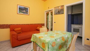 A room with a sofa, a table with a floral tablecloth, a kitchen area with blue tiles, and a door with glass panels. - M116 - Marcelli, two-room apartment in the center and facing the sea (Marcelli)