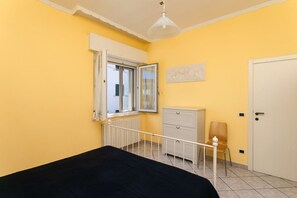 A bedroom with a bed, a radiator, a window, a white dresser, a chair, and a door.