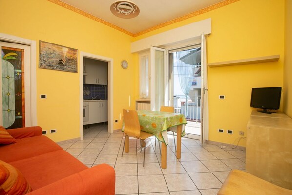 A bright yellow room with a dining table, chairs, a sofa, a TV, and a kitchen in the background.