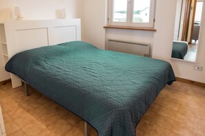 A single bed with a teal bedspread in a room with a white dresser, a window, and a mirror.