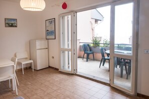 Una habitación con una mesa de comedor, sillas, un refrigerador y un ventanal corredizo que da a un balcón exterior con sillas.