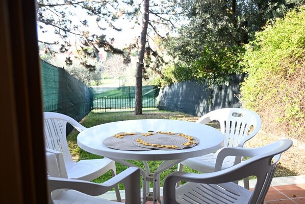 A round table with two white plastic chairs, surrounded by greenery and a fence.