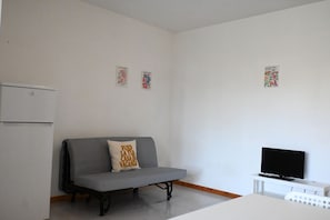 A small, clean room with a grey sofa, a flat-screen TV, and a refrigerator.