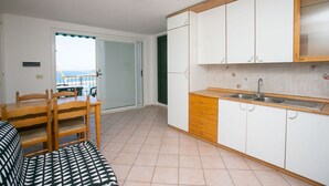 Cuina compacta amb armaris blancs, taulell de fusta i un rentaplat. Zona de menjador amb taula i cadires, i una vista per una porta.