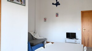Une pièce avec une table blanche, un canapé gris, un téléviseur posé sur un meuble blanc, un lustre et une porte en bois.