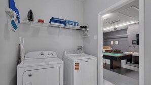 A laundry area with two washers and dryers, a shelf with folded towels, and a billiards table in the adjacent room.