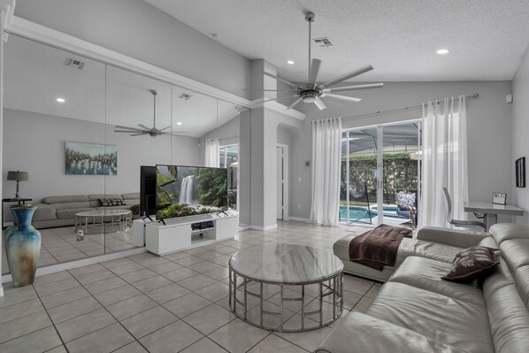 A spacious living room with a large flat-screen TV, a ceiling fan, and a view of an outdoor pool.