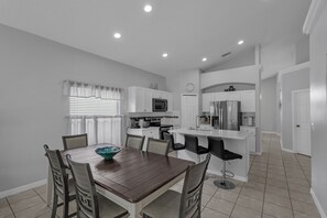 A modern kitchen with white cabinets, a central island with bar stools, a dining table with chairs, and a window with blinds.