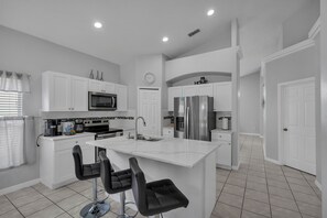 A modern kitchen with white cabinetry, a central island with bar stools, stainless steel appliances, and a clock on the wall.
