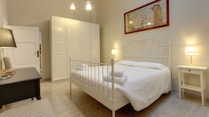 A bedroom with a white bed, a desk, a chair, a wardrobe, and a painting on the wall.