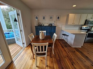Un coin repas avec une table en bois et des chaises blanches, un buffet bleu agrémenté d’objets décoratifs, et une cuisine avec des meubles blancs et des appareils électroménagers.
