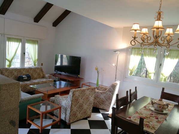 A living room with a flat-screen TV, floral-patterned sofas, a dining table with chairs, and a chandelier.