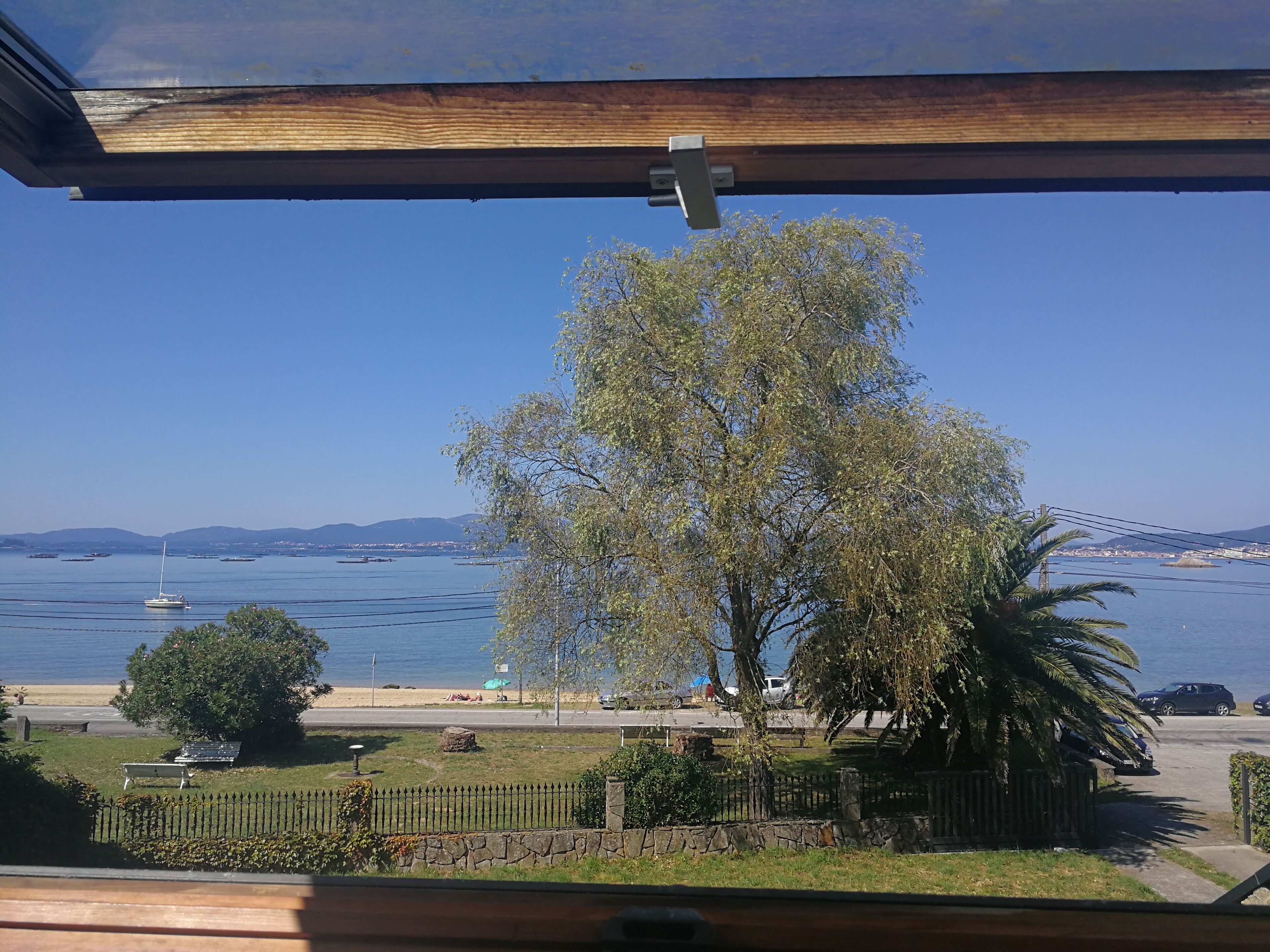 A view from a window overlooking a beach with a sailboat, trees, and a distant bridge.