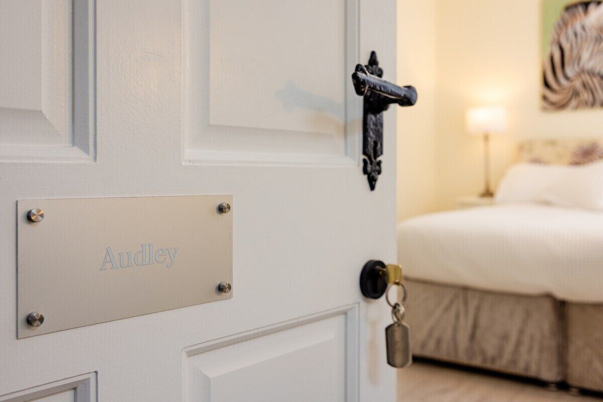 A hotel room with a white door, a nameplate reading "Audley," a black door handle, and a bed with a white comforter.