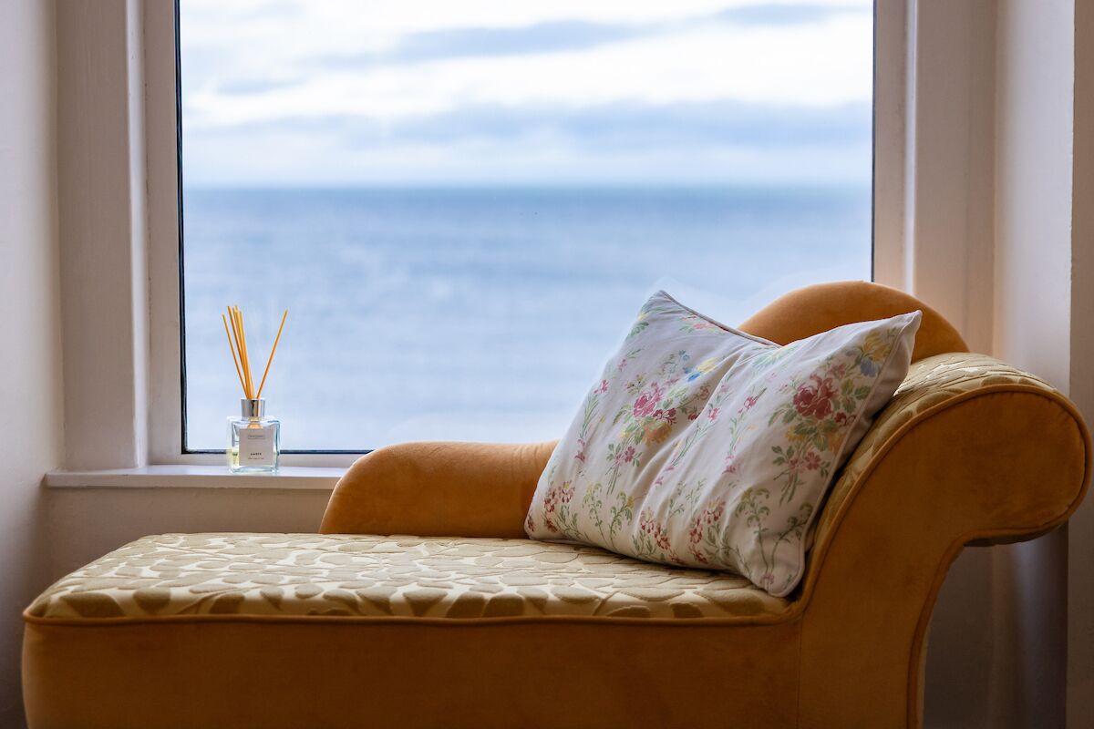A room with a view of the sea through a window, a sofa with a floral pillow, and a bottle of reed diffuser.