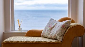 A room with a view of the sea through a window, a sofa with a floral pillow, and a bottle of reed diffuser.