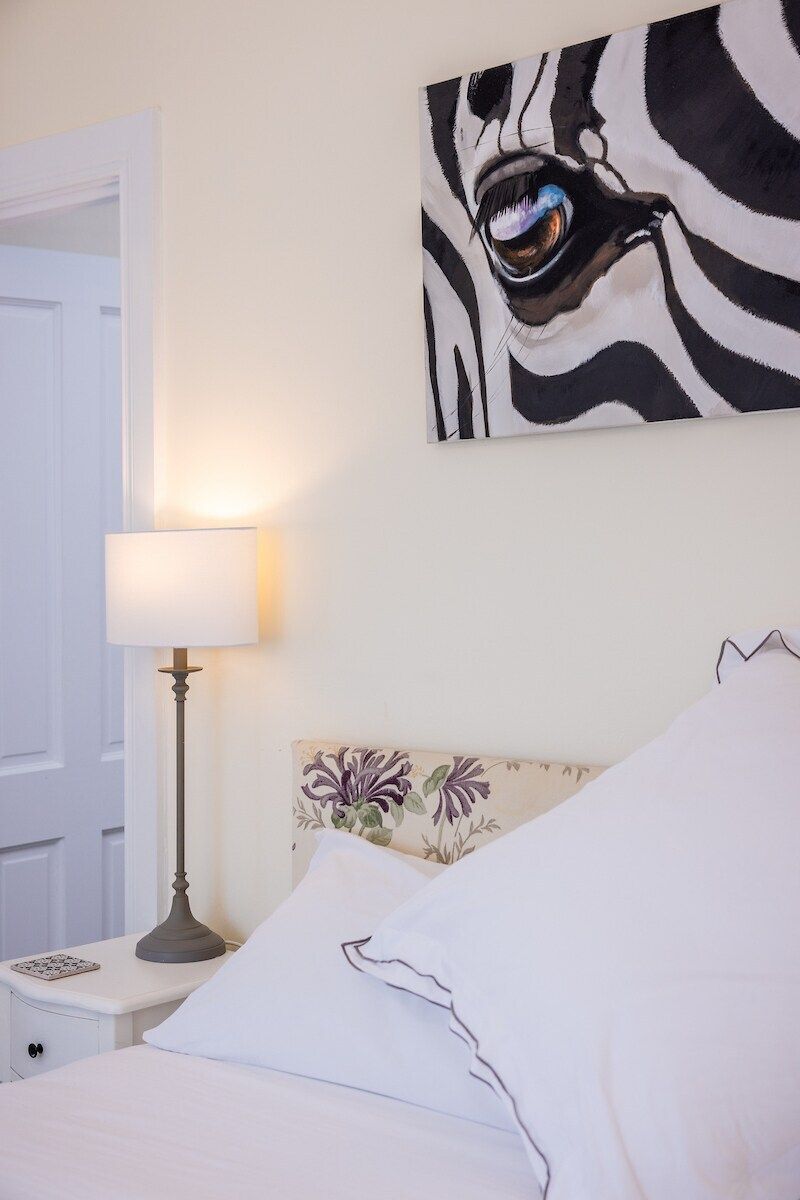 Un dormitorio con una cama, una mesita de noche con una lámpara, un cuadro de cebra en la pared y un cabecero con motivos florales.