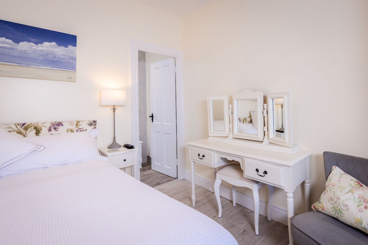 A bedroom with a bed, a vanity with mirrors, a lamp, and a door leading to another room.
