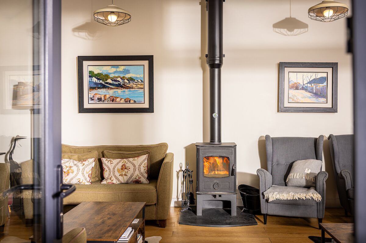 A cozy living room with a wood-burning stove, a sofa, armchairs, and two framed paintings on the wall.