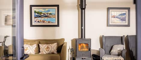 A cozy living room with a wood-burning stove, a sofa, armchairs, and two framed paintings on the wall.