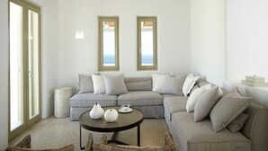 Ein geräumiges Wohnzimmer mit einer großen Eckcouch, einem runden Couchtisch mit dekorativen Vasen und einem Deckenventilator. Zwei Fenster bieten Blick auf das Meer.