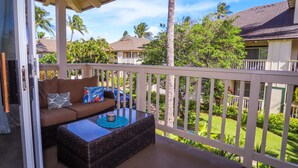 A balcony with a wicker sofa, a glass-top coffee table, and a view of a tropical neighborhood with palm trees and houses. - 3BR condo with A/C, pool & 10min walk to the beach! (Koloa)