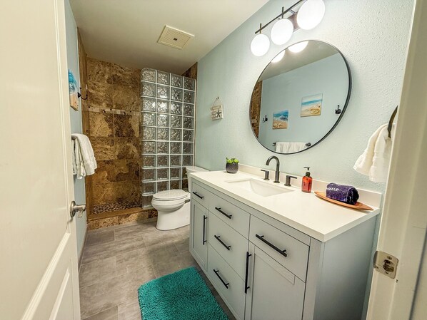 A bathroom with a stone shower, a white toilet, a sink with a mirror, and a teal rug.