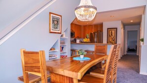 A dining area with a wooden table and chairs, a chandelier, and a kitchen in the background. - 3BR condo with A/C, pool & 10min walk to the beach! (Koloa)