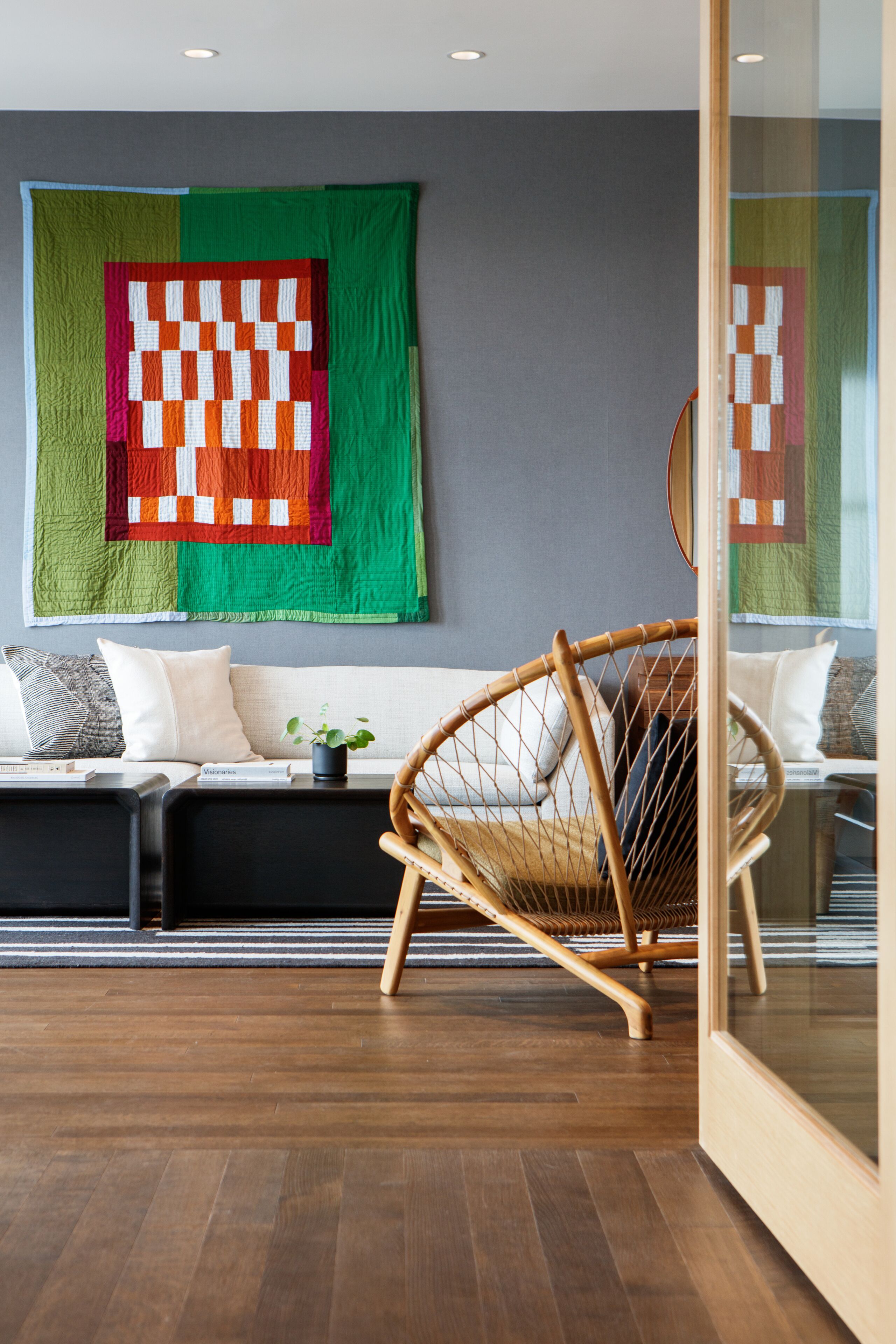 A modern living room with a wooden floor, a wicker chair, a sofa with cushions, and a large quilt hanging on the wall.