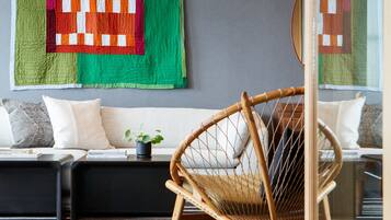 A modern living room with a wooden floor, a wicker chair, a sofa with cushions, and a large quilt hanging on the wall.
